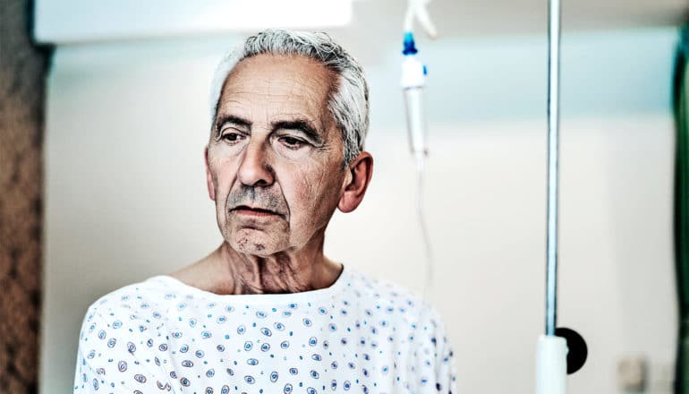 An older man sits on a hospital bed in a gown with an IV drip next to him