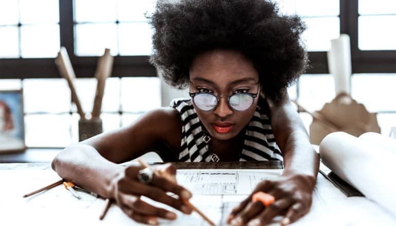 A woman with glasses and a striped shirt looks focused as she looks down at blueprints while holding a pencil