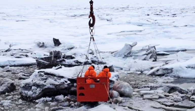 Researchers in orange cold-weather gear stand in a red box being lowered by a winch over a huge plain of sea ice