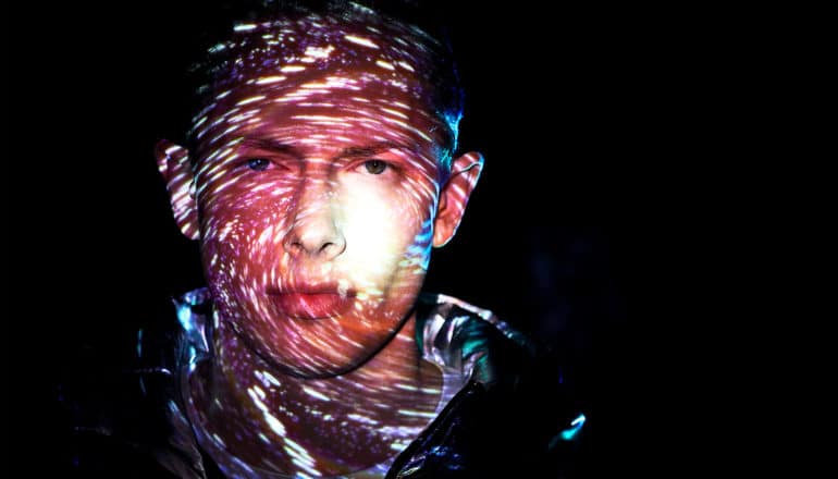 A purple-colored projection of swirling stars and space light up a man's face as he stands against a black background