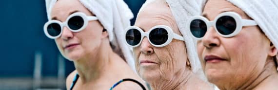 Three women in the same blue bathing suits, white sunglasses, and white towels wrapped around their heads sit by the pool, the youngest in the rear