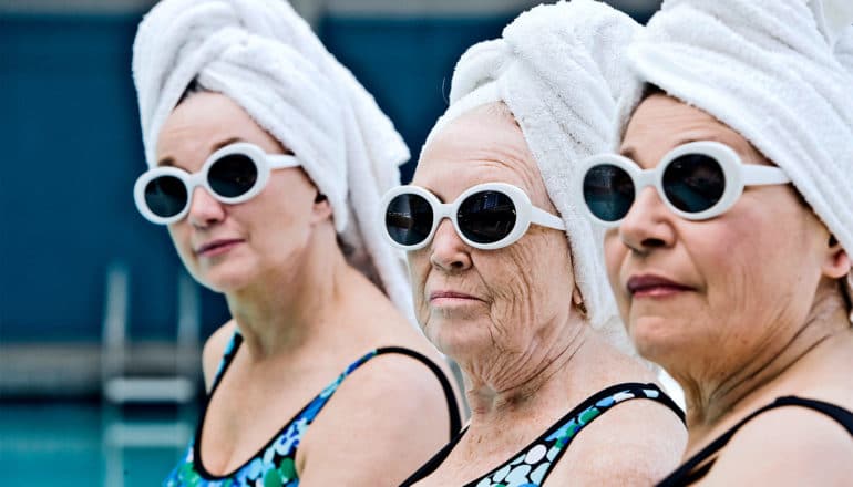 Three women in the same blue bathing suits, white sunglasses, and white towels wrapped around their heads sit by the pool, the youngest in the rear