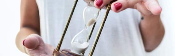 A woman in a white t-shirt turns an hourglass over in her hands, with her nails painted red
