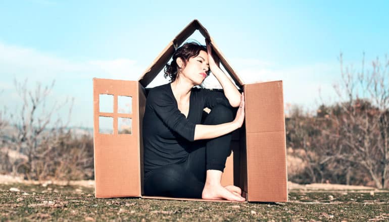 A woman sits looking anxious in a box cut to look like a tiny house