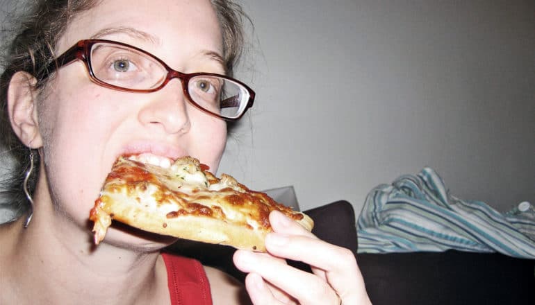 A young woman eats a slice of pizza while in her bedroom