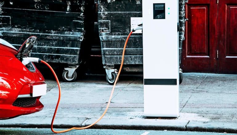 A red electric car charges parked at a curb, with a cord running into its trunk from an electric charging station