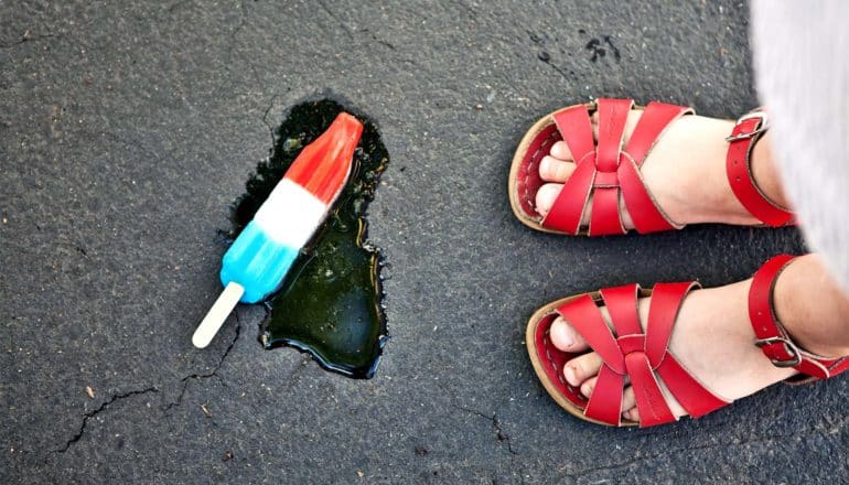 A young girl has dropped a red, white, and blue popsicle on concrete. The popsicle melts near her red sandals