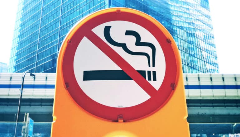 A yellow no-smoking sign stands on a street in front of a large skyscraper in the background