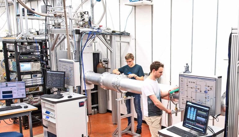 two people in lab with silver tube, wires, and computer equipment