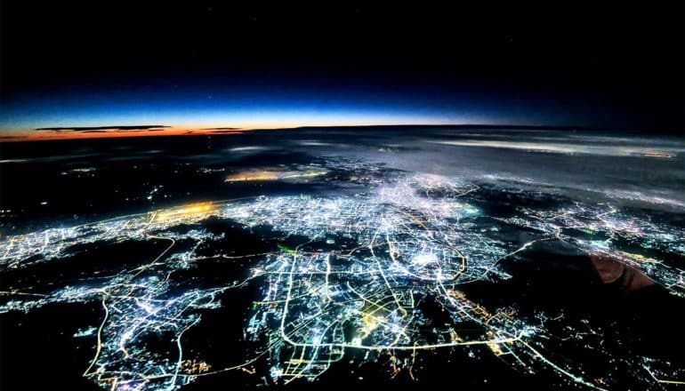 Nighttime city lights stretch over the horizon with the sunset in the distance over the Earth's curve