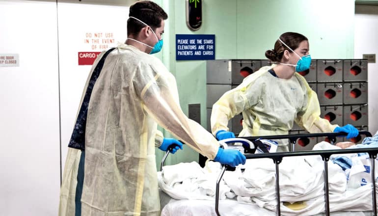 Two sailors in protective masks, gloves, and gowns wheel a gurney with a patient on it down a hallway