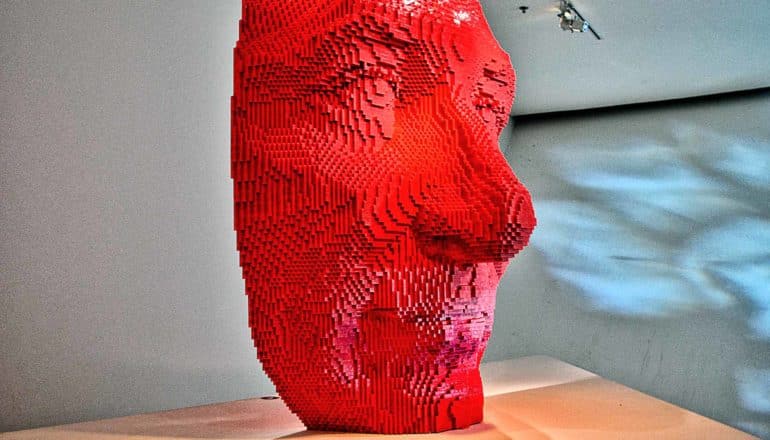 A face made of red lego bricks sits on a wooden pedestal at an art gallery