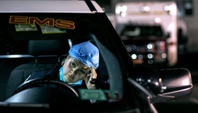 person in vehicle with PPE hair net has sad expression, puts hand to temple