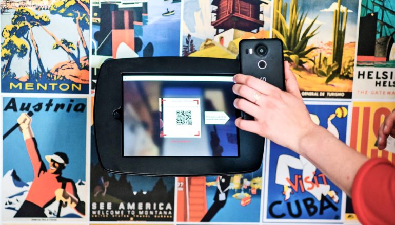 hand holds smartphone up to screen displaying QR code. Background of travel posters