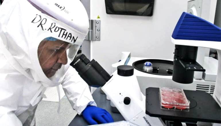 A researcher wearing a full protective suit looks through a microscope