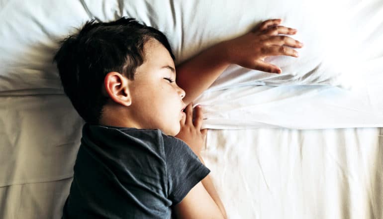 A young boy sleeps on his side on white sheets in a gray t-shirt, with his left arm sticking out from under his face