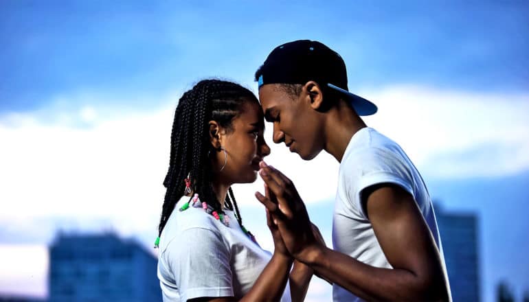 Two teens touch hands and foreheads with a city and blue sky in the background