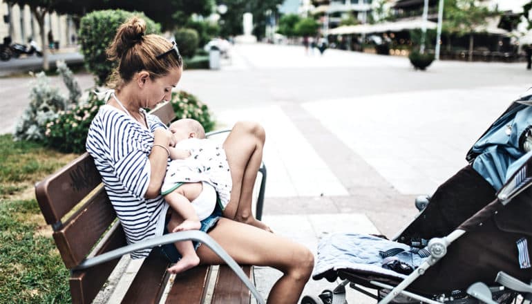 A mom breastfeeds her baby on a bench outside