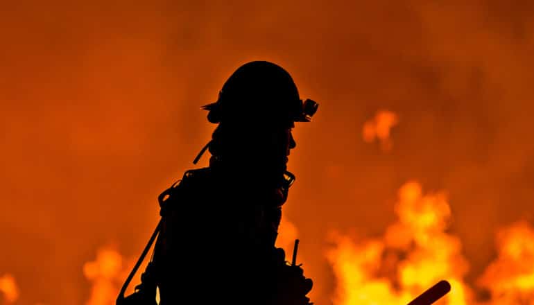 A firefighter walks past a wildfire which is casting orange light behind him