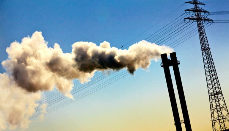 A smoke stack in front of a power line spews a cloud into the blue sky