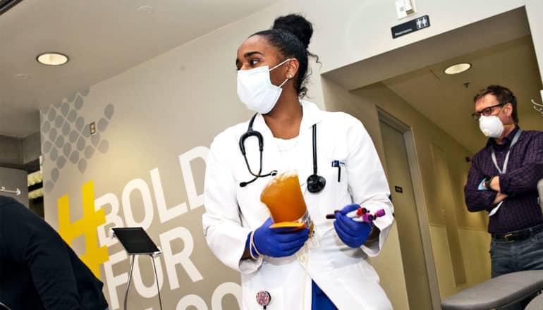 A phlebotomist in a white coat carries a yellowish bag of convalescent plasma from a COVID-19 donor