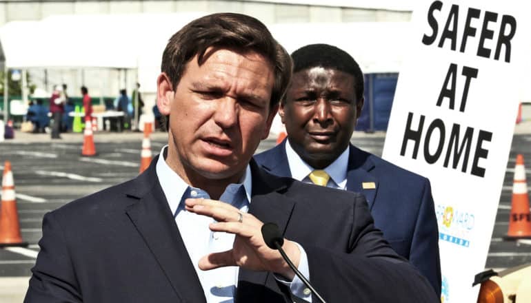 Governor Ron DeSantis speaks in a parking lot in front of a sign that reads "Safer at Home"