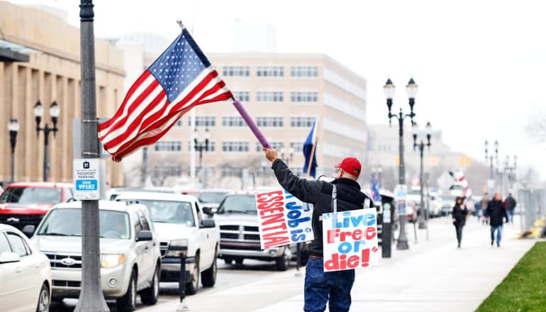 A protestor holds an American flag and two signs, one that reads "Live Free or Die!" and another that reads "A job is essential"