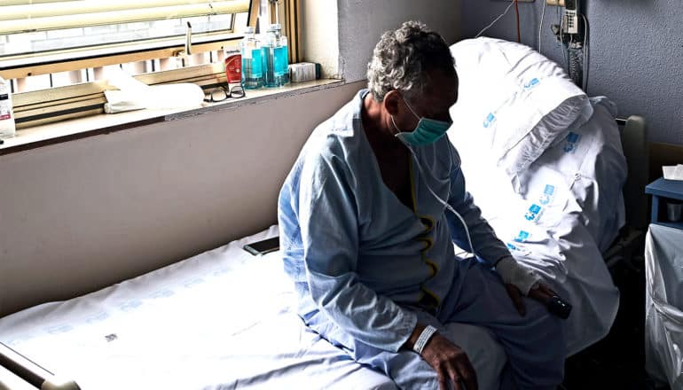 A man in pajamas and a medical mask, obscured by shadow, sits up at the edge of a hospital bed