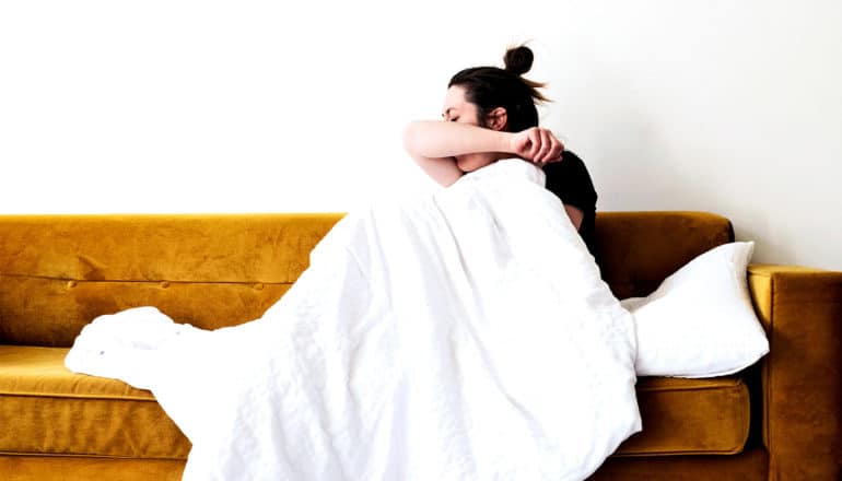 A woman coughs into her elbow while on her couch under a blanket