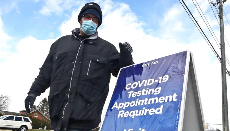 A man in winter clothes and a medical mask puts a blue sign that reads "COVID-19 Testing Appointment Required"