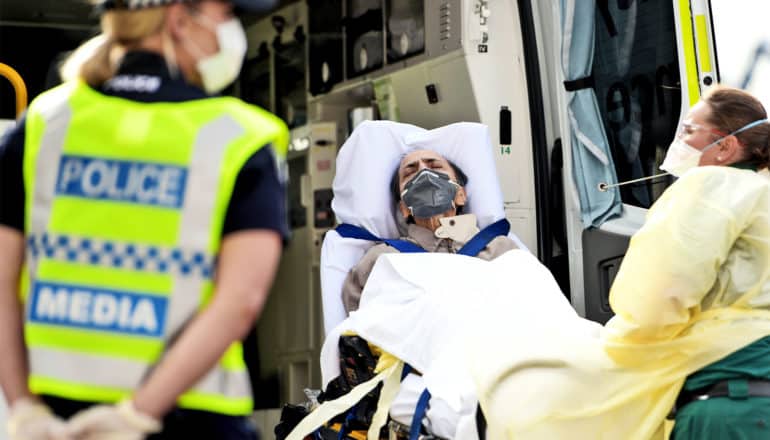 Health care workers put a man on a stretcher into an ambulance as a police officer with a mask on looks on