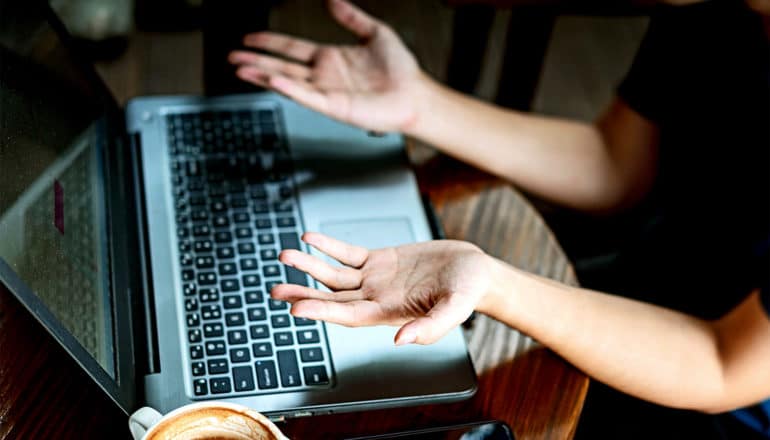 A person holds their hands up in exasperation while looking at their laptop