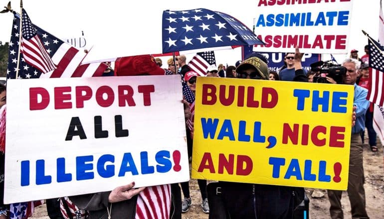 Protesters hold American flags and signs that read "Deport all illegals," "Build the wall, nice and tall," and "Assimilate, Assimilate, Assimilate"