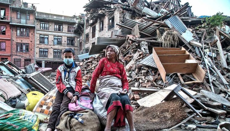 A grandmother and grandson sit on bags of belongings in front of a destroyed home