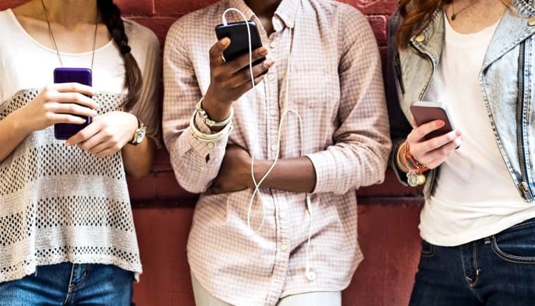 Three women look at their phones while standing against a wall