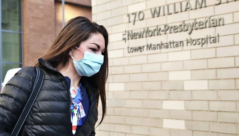 A nurse wearing a black coat and blue medical mask walks into work at a Manhattan hospital