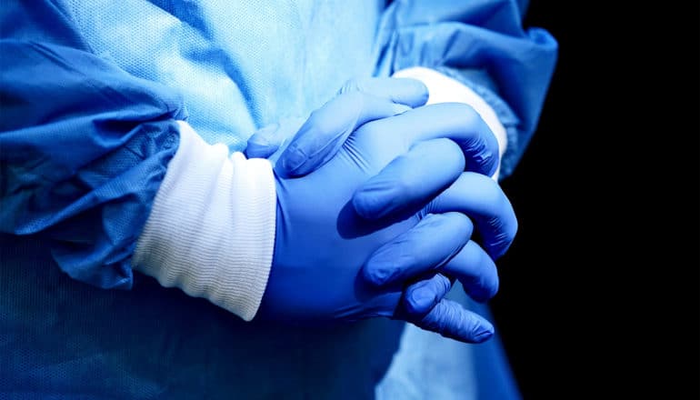 A health care worker clasps blue-gloved hands together