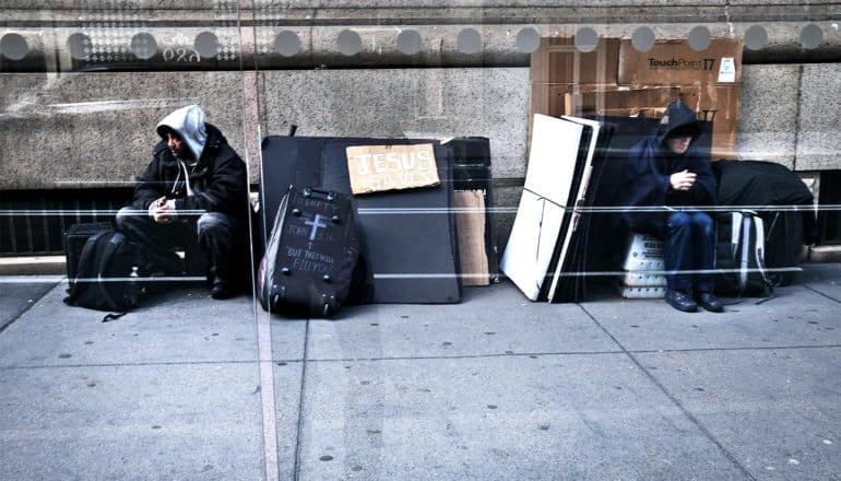 Two men sit on the sidewalk near a few belongings