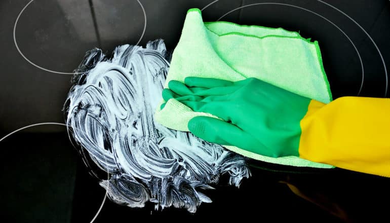 A hand with a dish glove on uses a green cloth to clean a black stove top