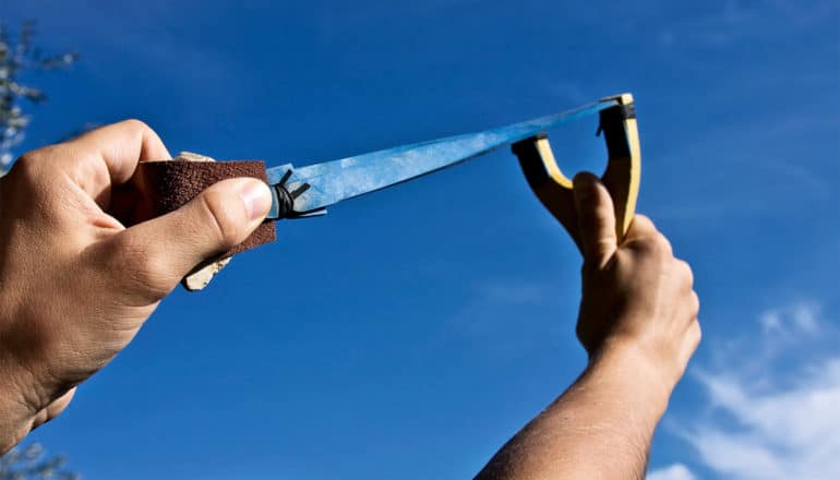 A man holds a slingshot pointed at the blue sky