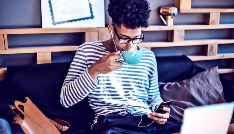 A young man drinks coffee while looking at his phone and listening to music