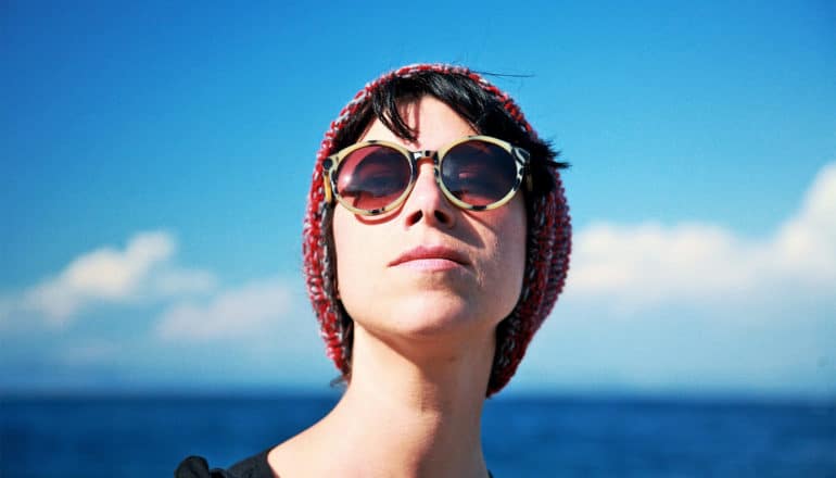 A young woman with a red beanie and red glasses stands outside looking up