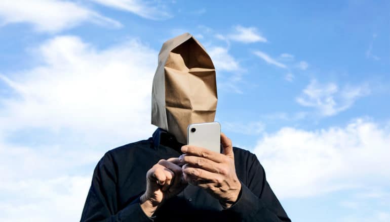 A man has a paper bag on his head as he looks at his phone, with blue sky in the background