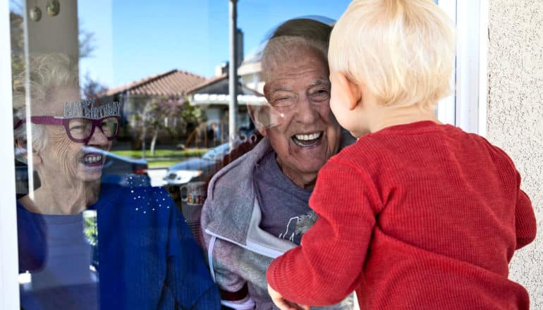 Two older adults greet a young boy in red through their window