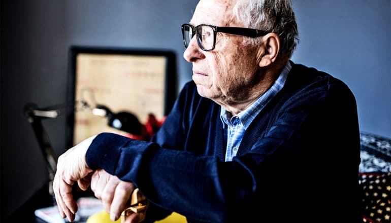 An older man in a navy sweater leans on a cane while he's sitting looking off into the middle distance