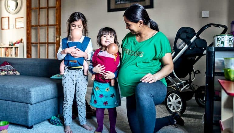 A pregnant mother watches her two daughters practice holding a baby in a bjorn