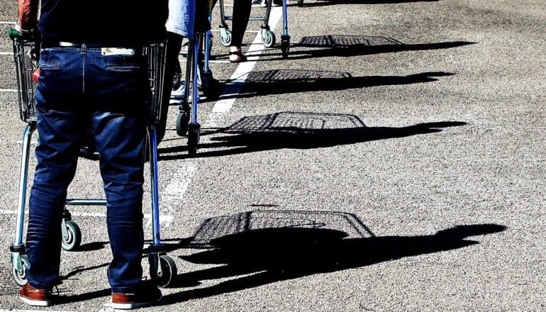 Grocery shoppers stand 6 feet apart in the parking lot with shopping carts, their shadows visible on the asphalt