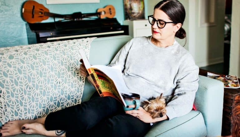 A woman reads a book while sitting on her couch with a cup of coffee and her dog