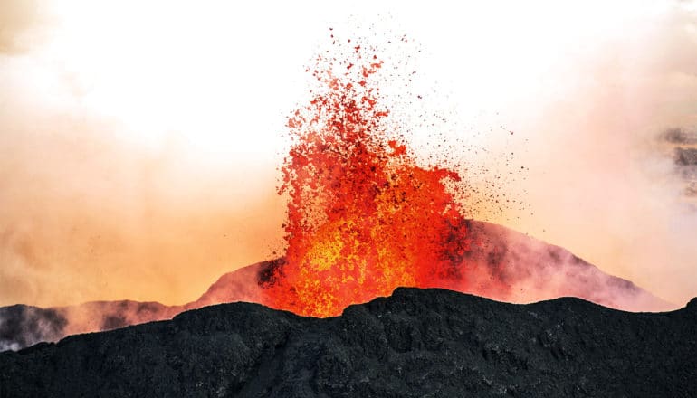 A volcano erupts, sending orange magma flying and making smoke around the eruption glow orange