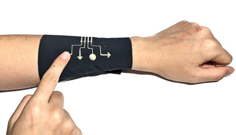 A researcher wears the black wearable electronic fabric on his arm. It has several designs that look like directional buttons